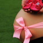 Pregnant woman with bare belly bandaged with pink ribbon holding pink booties